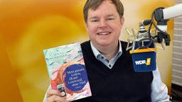 Mike Altwiker hält das Buchcover "Mein ganzes Leben, Öl auf Leinwand, ohne Titel" von Alena Schröder in die Kamera