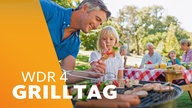 Ein Vater steht mit seiner kleinen Tochter am Grill und erklärt. Im Hintergrund weitere Menschen an einem gedeckten Picknicktisch. Sonniger Tag, grüne Umgebung.