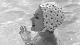 Schwarz-weiße Aufnahme einer jungen Frau im Schwimmbad mit einer für die 1960er Jahre typischen Badekappe aus einem mit Blumen gemusterten Kunststoff