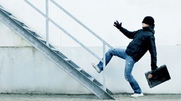 Ein dunkel gekleideter Bankräuber mit schwarzer, tief heruntergezogener Mütze und einem Koffer in der Hand läuft auf der Flucht eine Treppe hoch. 