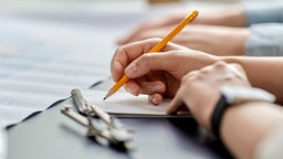 Eine Hand schreibt mit einem orangenen Bleistift auf einem Notizblock. An der anderen Hand trägt die Person eine Uhr, auf dem Tisch neben dem Notizblock liegt eine Brille. 