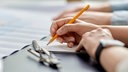 Eine Hand schreibt mit einem orangenen Bleistift auf einem Notizblock. An der anderen Hand trägt die Person eine Uhr, auf dem Tisch neben dem Notizblock liegt eine Brille. 