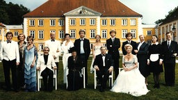 Bild zu der Serie "Das Erbe der Guldenburgs": Die Schauspieler stehen schick gekleidet in einer Reihe vor einem gelben Schloss