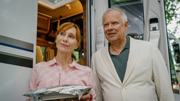 Gundula (Andrea Sawatzki, l.) steht mit Gerald (Axel Milberg, r.) aus dem Film "Familie Bundschuh – Wir machen Camping" mit Essen in der Hand vor einem Camper.
