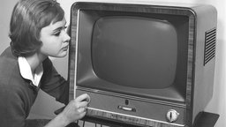 Eine junge Frau ist neben einem typischen Fernsehgerät der 50er Jahre abgebildet