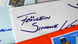 Ein Briefumschlag, auf der mit blauer Tinte "Fräulein Simone" steht. Der Umschlag ist teilweise mit einem anderen Briefumschlag abgedeckt, weitere Briefumschläge liegen darunter und drumherum. 