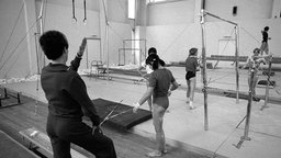 Sechs Personen stehen verteilt in einer Turnhalle, in der einige Geräte zum Turnen aufgebaut sind
