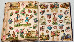 Ein aufgeschlagenes Poesiealbum, dessen Seiten mit vielen kleinen Glanzbildern beklebt sind. Bei den Motiven handelt es sich vor allem um Herzen und Blumen, auch einige Glanzbilder mit Ostereiern sind zu sehen. 