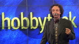 Moderator Jean Pütz vor dem Schriftzug "Hobbythek", am gestikulieren. Er trägt ein graues Sakko, darunter eine gemusterte Weste und ein graues Hemd sowie eine dunkelrote Fliege. 