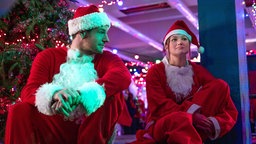 Eine Szene des Films "Jingle Bell Heist": Nick, gespielt von Connor Swindells, und Sophia, gespielt von Olivia Holt, sitzen in Weihnachtsmannkostümen auf dem Rand einer Tribüne. Die Kostümbärte haben sie heruntergezogen, er schaut sie interessiert an.
