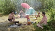 Ein Foto aus der Ausstellung: Im Vordergrund hockt ein Mann in Badehose an einem Campinggrill und grillt, ein weiterer Mann sitzt daneben im Gras und schaut ihm zu. Im Hintergrund sitzt eine Frau im Badeanzug vor einem Zelt unter einem Sonnenschirm.