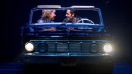 Darsteller Antonia Kalinowski und Philipp Büttner als Sandy und Danny auf der Bühne. Sie sitzen in einem Auto, er am Steuer, sie auf dem Beifahrersitz, und schauen einander lächelnd an.
