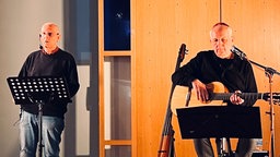 Autor Holger Jenrich steht mit einem Headset-Mikrofon auf der Bühne. Musiker Lutz Debus sitzt mit etwas Abstand rechts neben ihm. Er spielt Gitarre, vor ihm ein Mikrofon und ein Notenständer. Er trägt eine Kippa. 