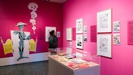 Ein Ausstellungsraum mit pinkfarbenen Wänden: Eine Besucherin schaut sich einen der Comics an, die in Rahmen an den Wänden hängen. Im Vordergrund ist ein Ausstellungskasten aus Glas mit "Lucky Luke"-Ausgaben zu sehen. 