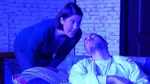 Schauspieler auf einer Bühne: Eine Frau beugt sich über einen Mann, der mit geschlossenen Augen auf einem Stuhl sitzt.