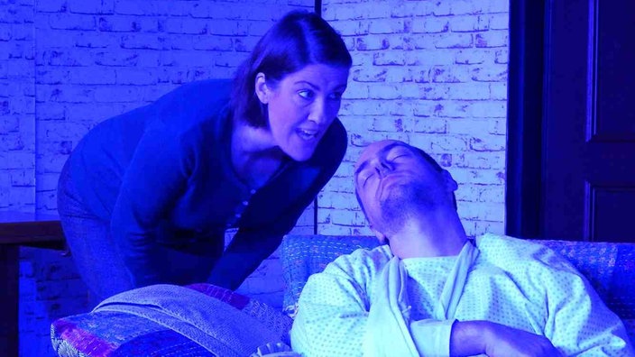Schauspieler auf einer Bühne: Eine Frau beugt sich über einen Mann, der mit geschlossenen Augen auf einem Stuhl sitzt.
