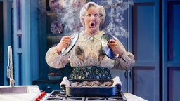 Ein Foto der Aufführung: Mrs Doubtfire steht mit schockiertem Gesichtsausdruck am Herd mit zwei dampfenden Topfdeckeln in der Hand, auf ihrer Bluse sind schwarze Rußflecken. 