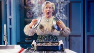 Ein Foto der Aufführung: Mrs Doubtfire steht mit schockiertem Gesichtsausdruck am Herd mit zwei dampfenden Topfdeckeln in der Hand, auf ihrer Bluse sind schwarze Rußflecken. 