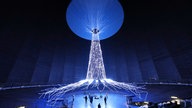 40 Meter hohe Lichtinstallation in Form eines Baums. Die Wurzeln breiten sich strahlenförmig aus. Darunter liegen Besucher der Ausstellung auf dem Boden und schauen den Baum hinauf nach oben. 