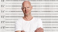 Rüdiger Hoffmann steht mit verschränkten Armen vor einer Polizei-Wand mit Messlatte