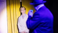 Eine Frau steht im Scheinwerferlicht eines Mannes, der auf sie leuchet. Die Frau trägt einen weißen Mantel und schaut verschreckt den Mann an. Der Mann trägt einen schwarzen Mantel und Hut.