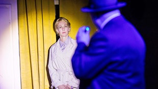 Eine Frau steht im Scheinwerferlicht eines Mannes, der auf sie leuchet. Die Frau trägt einen weißen Mantel und schaut verschreckt den Mann an. Der Mann trägt einen schwarzen Mantel und Hut.