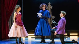 Julian Culemann als Michael, der als Frau verkleidet ist und sich Dorothy nennt steht in der Mitte der Bühne mit einem Skript in der Hand. Rechts und links von ihm befinden sich weitere Schauspieler.