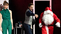 Eine Aufnahme aus der Aufführung: Darsteller Tina Seidel und Thomas Jansen sind festlich gekleidet. Jansen zeigt auf einen weiteren Darsteller, der als Weihnachtsmann verkleidet ist. Er hat die Kapuze tief in die Stirn gezogen und schleicht sich davon.