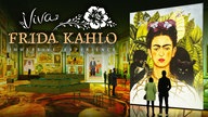 Das Bild zeigt eine farbenfrohe Ausstellung mit vielen Gemälden Frida Kahlos. Im Vordergrund rechts im Bild ist ein Selbstporträt von ihr zu sehen, davor stehen einige Besucher. Die Aufschrift "Viva Frida Kahlo, immersive Experience" ist außerdem zu sehen