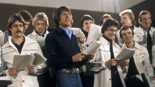 Die Fußballnationalmannschaft 1978 mit Udo Jürgens bei Aufnahmen zum WM-Song "Buenos Dias Argentina". Die Nationalspieler stehen in Trainingsjacken um Udo Jürgens herum, alle halten Textzettel in der Hand und singen. 