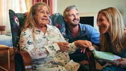 Filmszene aus dem Film "Noch einmal, June": June (Noni Hazlehurst) sitzt in einem grünen Sessel mit Blumenmuster. Rechts neben ihr befinden sich ihr Sohn Devon (Stephen Curry) und ihre Tochter Ginny (Claudia Karvan). June hält die Hand ihrer Tochter.