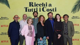 Ein Gruppenfoto des Casts des Films "Reich um jeden Preis" bei der Premiere auf dem roten Teppich. Die acht Schauspieler lächeln freundlich in die Kamera. Hinter ihnen an der gelben Wand steht der italienische Originaltitel "Ricchi a Tutti i costi".