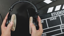 Eine Filmklappe liegt auf einem schwarzen Untergrund, davor wird ein Paar Kopfhörer gehalten.