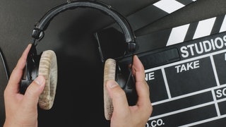 Eine Filmklappe liegt auf einem schwarzen Untergrund, davor wird ein Paar Kopfhörer gehalten.