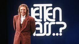 Der junge Thomas Gottschalk steht vor einem "Wetten dass..?" Schild und lächelt in die Kamera.