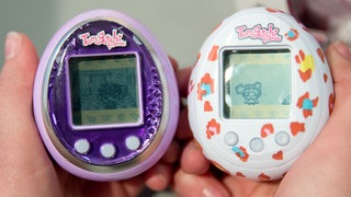 Ein lilafarbenes und ein weißés Tamagotchi werden nebeneinander gehalten.