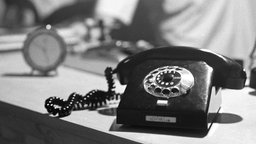 Schwarz-weiß-Foto eines Telefons mit Wählscheibe und einer Uhr im Hintergrund