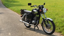 Ein altes Motorrad mit Zweitaktmotor, rundem Scheinwerfer und einfacher Sitzbank, welches auf einem Weg neben einer Wiese abgestellt ist.