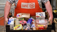 Eine Frau mit einer orangenen Schürze der Tafel präsentiert eine Kiste mit Lebensmitteln.
