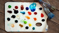 Eine Farbpalette mit zahlreichen bunten Acrylfarbtupfern liegt zusammen mit zwei Pinseln auf einem Holzboden
