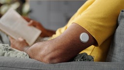 Ein kleiner, weißer, runder Glukosemonitor am Oberarm eines Mannes. Er trägt ein gelbes T-Shirt und sitzt lesend auf dem Sofa.