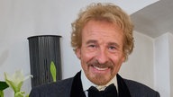 Thomas Gottschalk im schwarzen Anzug mit schwarzer Fliege. Er lächelt in die Kamera. 