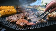 Jemand wendet die Würstchen, die mit Fleisch und Maiskolben auf einem Grill liegen und brutzeln