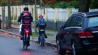 Eine Radfahrerin mit einem Kind im Fahrradsitz, daneben ein etwas älteres Kind auf einem eigenen Fahrrad. Das ältere Kind trägt einen Schulranzen auf dem Rücken. Alle tragen Helme. Sie fahren auf der Straße, am Straßenrand ist ein Auto geparkt. 