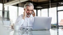 Ein Mann in einem weißen Hemd sitzt am Arbeitstisch vor einem Laptop und hält sich die schmerzenden Schläfen