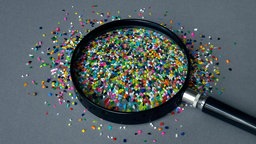 Eine Lupe liegt auf vielen, kleinen, bunten Plastikstückchen 