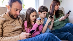 Vater, zwei Kinder, Mutter sitzen nebeneinander auf dem Sofa und starren auf ihre Smarthphones