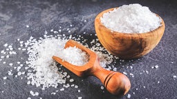 Grobkörniges Salz in einer Holzschale. Daneben eine kleine Holzschaufel mit Salz, weiteres Salz herum verstreut auf dem dunklen Steinuntergrund.