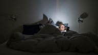Eine Frau liegt im Bett und bedient ihr Smartphone. Bis auf das Licht vom Display ist das Schlafzimmer dunkel. 