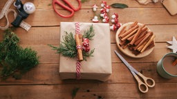 Ein Weihnachtsgeschenk in braunem Packpapier, verziert mit Tannengrün und Zimtstangen. Um das Paket herum auf dem braunen Holztisch liegen die Materialien, die zum Verpacken verwendet wurden. Am rechten Rand des Bildes steht außerdem eine Tasse Kaffee.